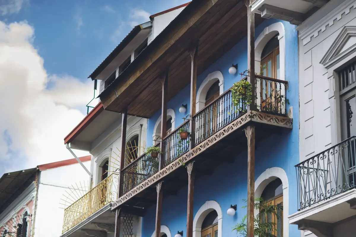 Balcón corrido de madera calada con tejadillo, pies derechos y barandilla de hierro con macetas, en la fachada azul de un edificio antiguo bajo un cielo con nubes