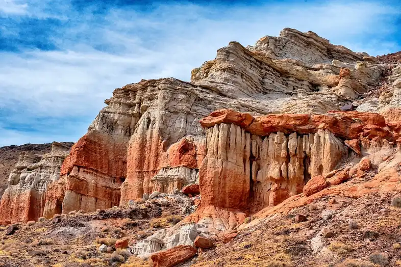 Formación rocosa erosionada en capas blancas y rojizas, con columnas verticales, sobre una ladera seca bajo un cielo azul con nubes