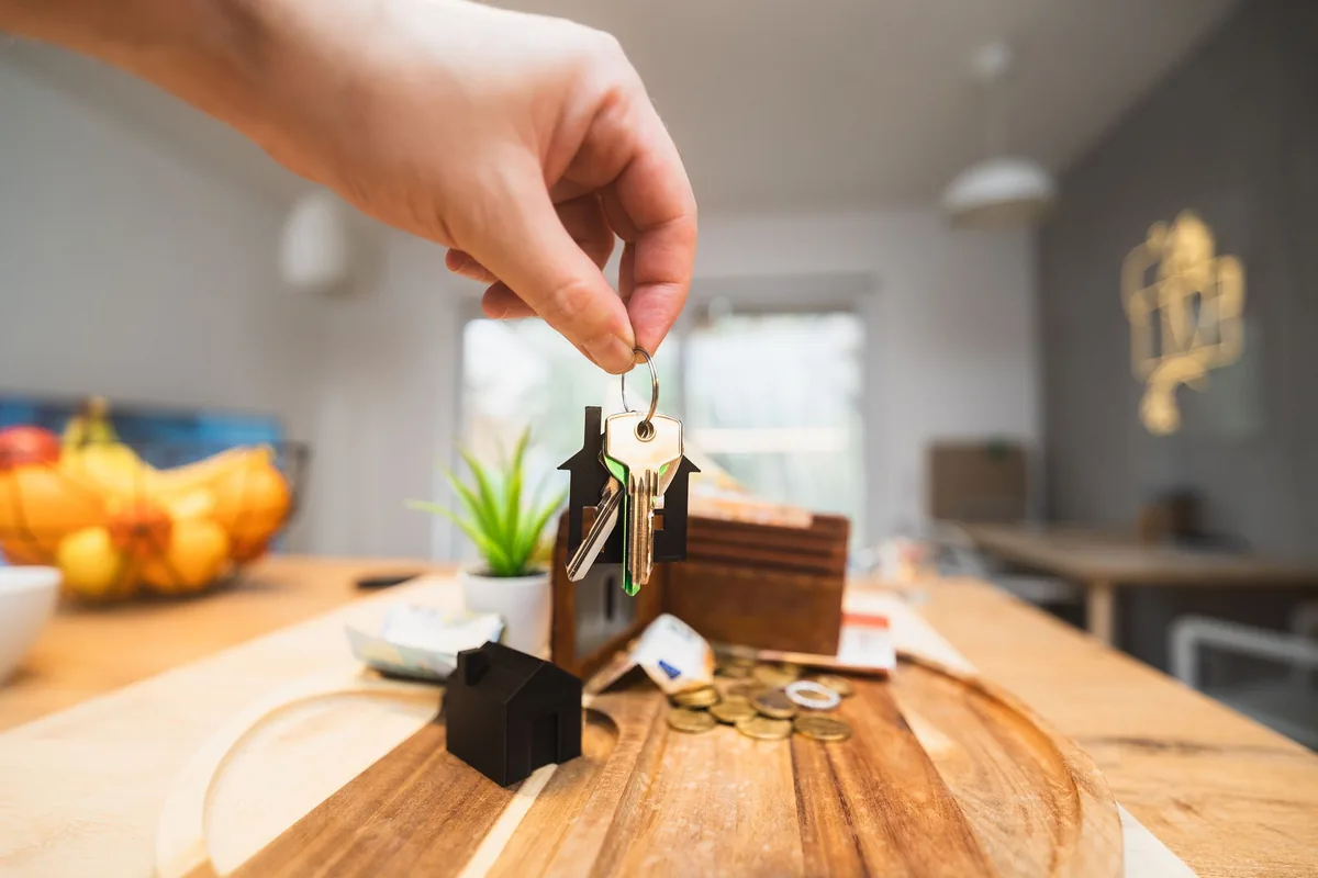 Mano con unas llaves de vivienda junto a casas en miniatura y monedas