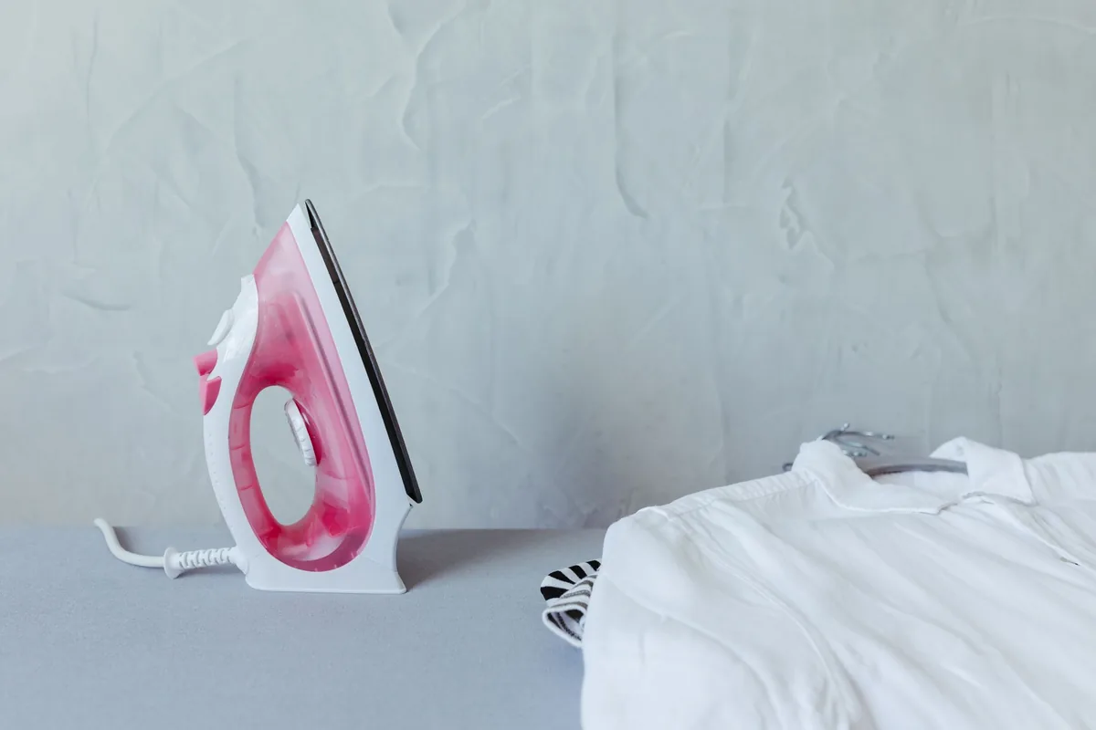 Plancha de vapor rosa y blanca sobre una tabla de planchar junto a una camisa blanca