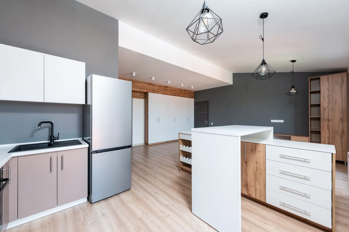 Cocina abierta de una vivienda con frigorífico plateado, isla blanca con cajones y lámparas colgantes de rejilla