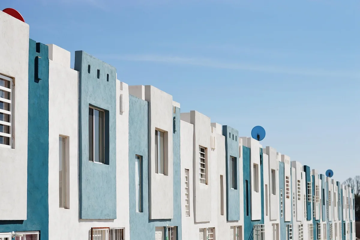Hilera de viviendas de dos plantas con fachadas blancas y azules, ventanas con rejas y antenas parabólicas bajo un cielo despejado