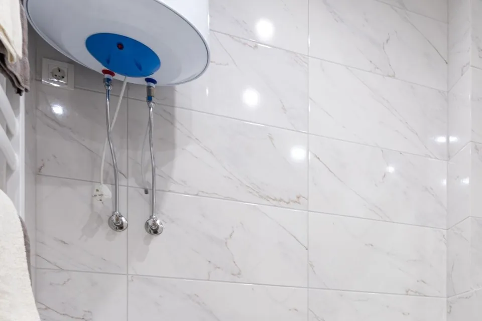 Termo eléctrico blanco con tapa azul colgado de una pared de azulejos, con dos tubos flexibles y un enchufe al lado