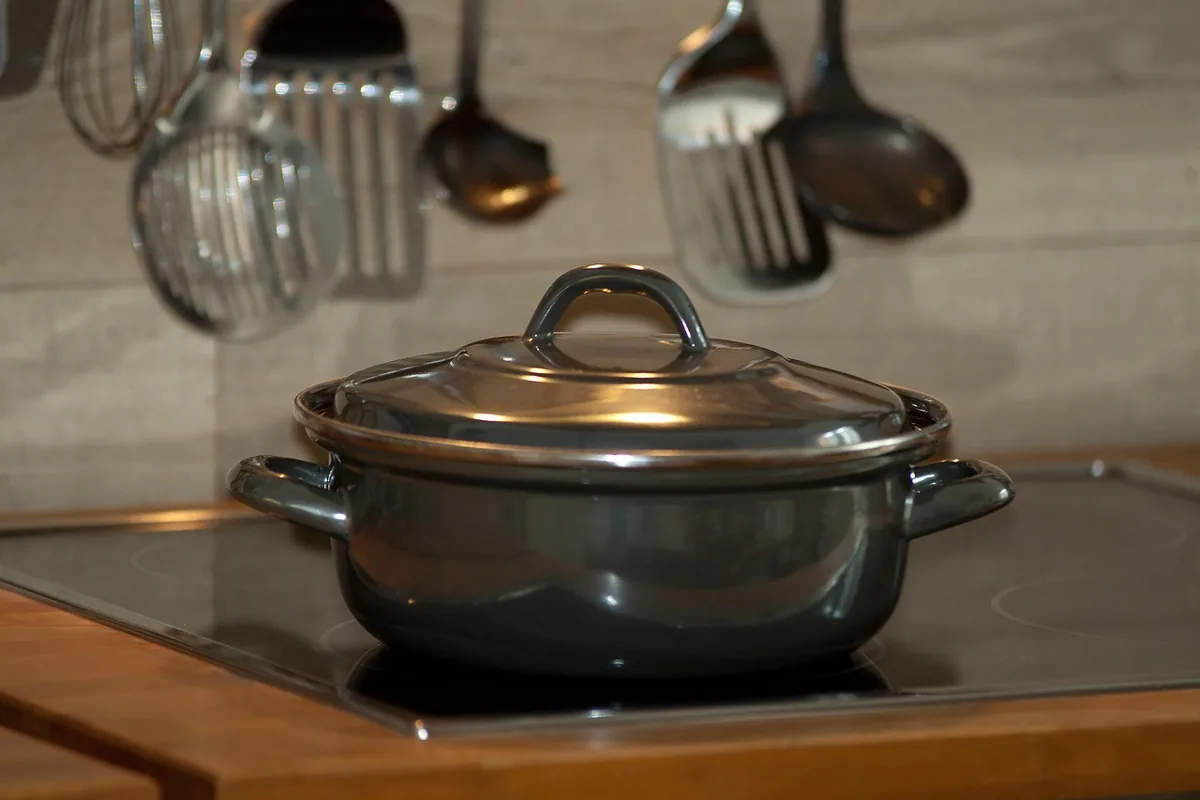 Cazo gris con tapa sobre una placa vitrocerámica, con utensilios de cocina colgados al fondo