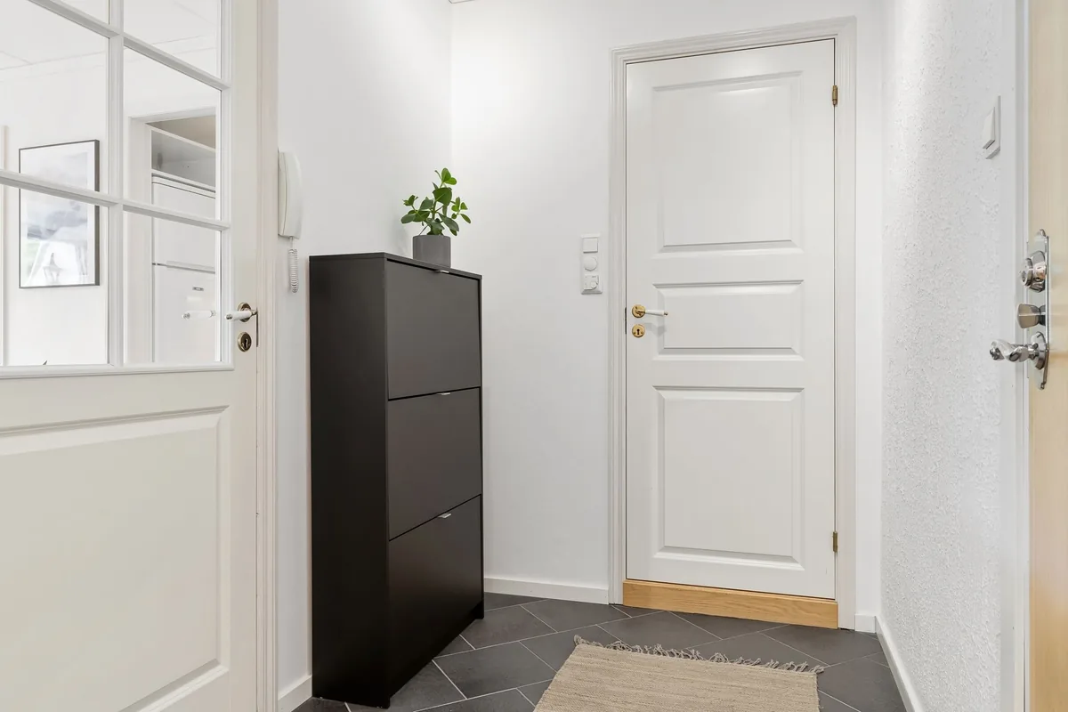 Recibidor de una vivienda con un mueble zapatero oscuro, una puerta blanca y, al fondo, una cocina con nevera