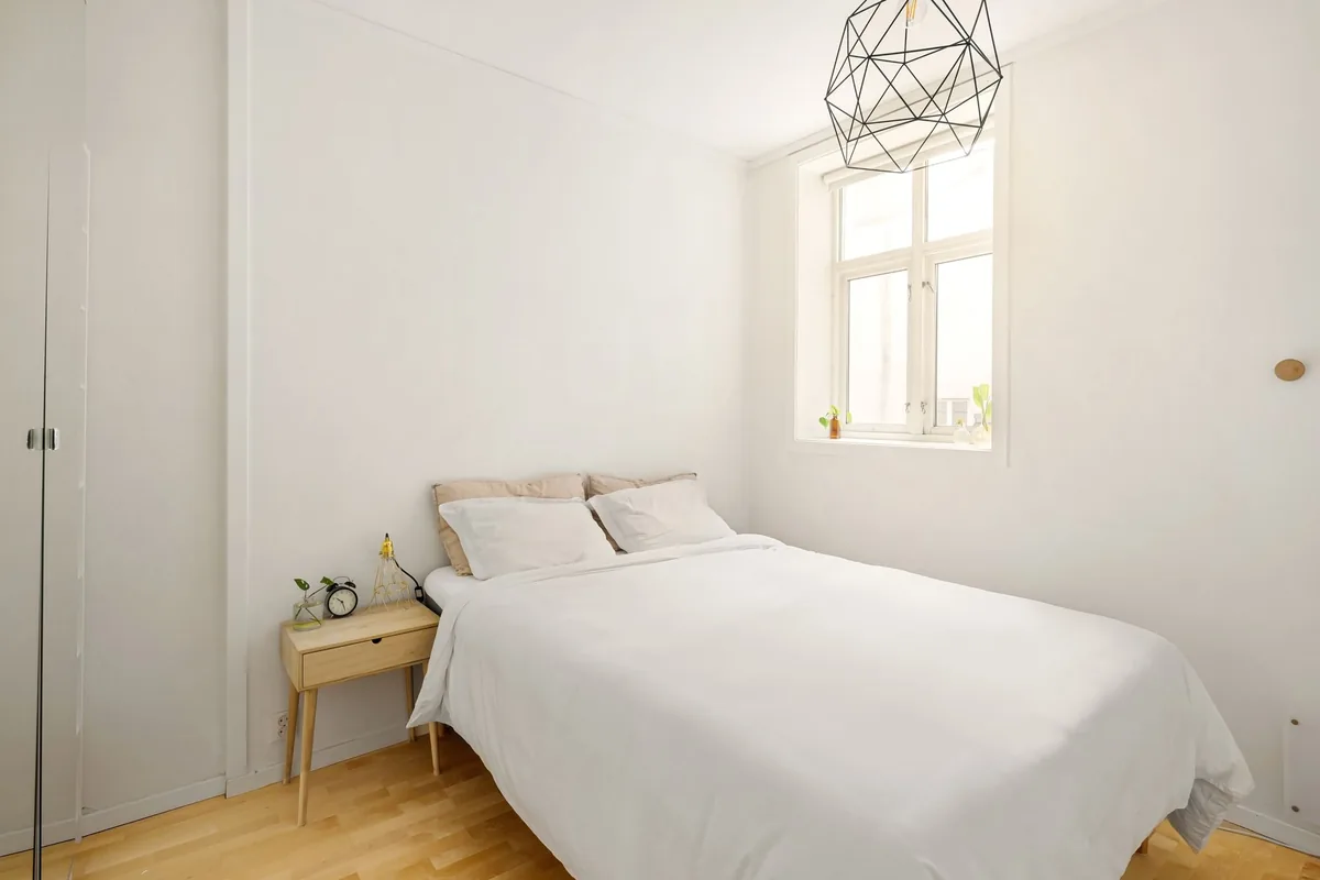 Dormitorio blanco con cama de matrimonio, mesilla de madera con un despertador y una lámpara de alambre en el techo