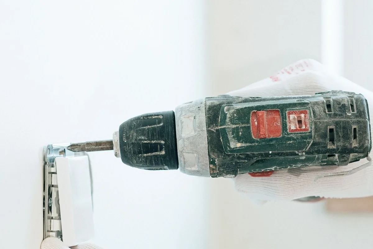 Manos con guantes atornillando con un taladro inalámbrico un mecanismo eléctrico en una pared blanca