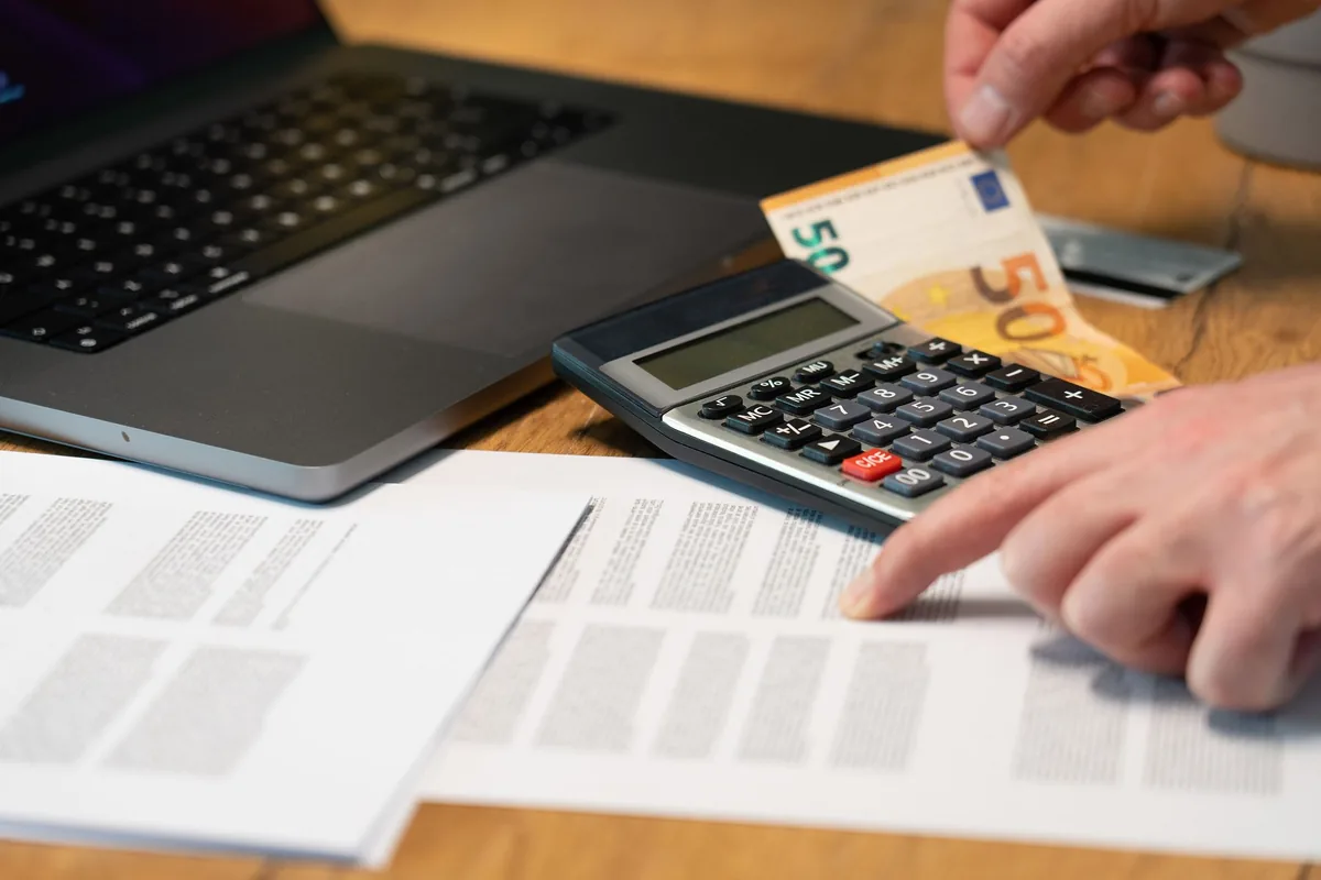 Mano junto a una calculadora, papeles, un portátil y un billete de 50 euros sobre una mesa de madera